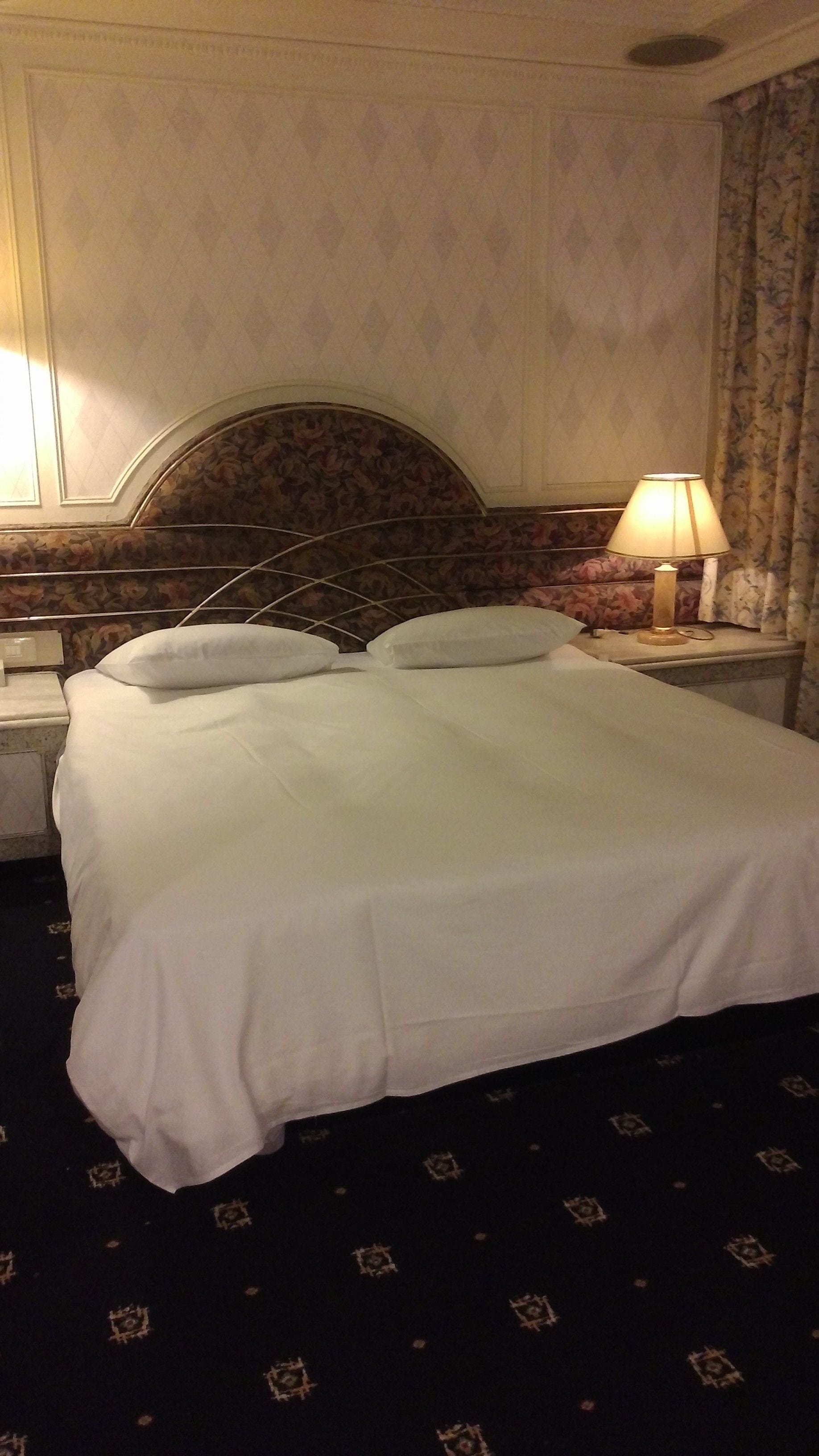 A hotel room with a large bed, a headboard with floral patterns, a nightstand with a lamp, and a patterned carpet.