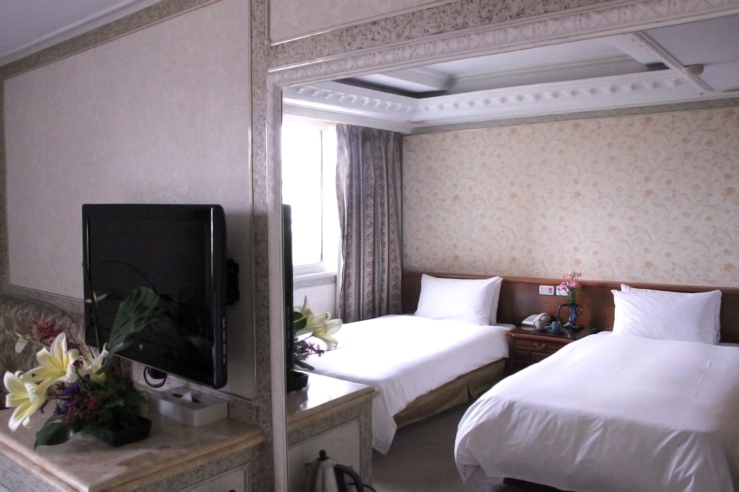 Ein Hotelzimmer mit zwei Betten, einem Fernseher, einem Schreibtisch mit Telefon und einem fenster mit Vorhängen.