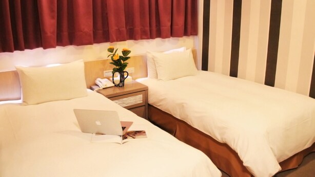 Quarto de hotel com duas camas, uma mesa de cabeceira com uma lâmpada e um vaso de flores, e um laptop em uma das camas.