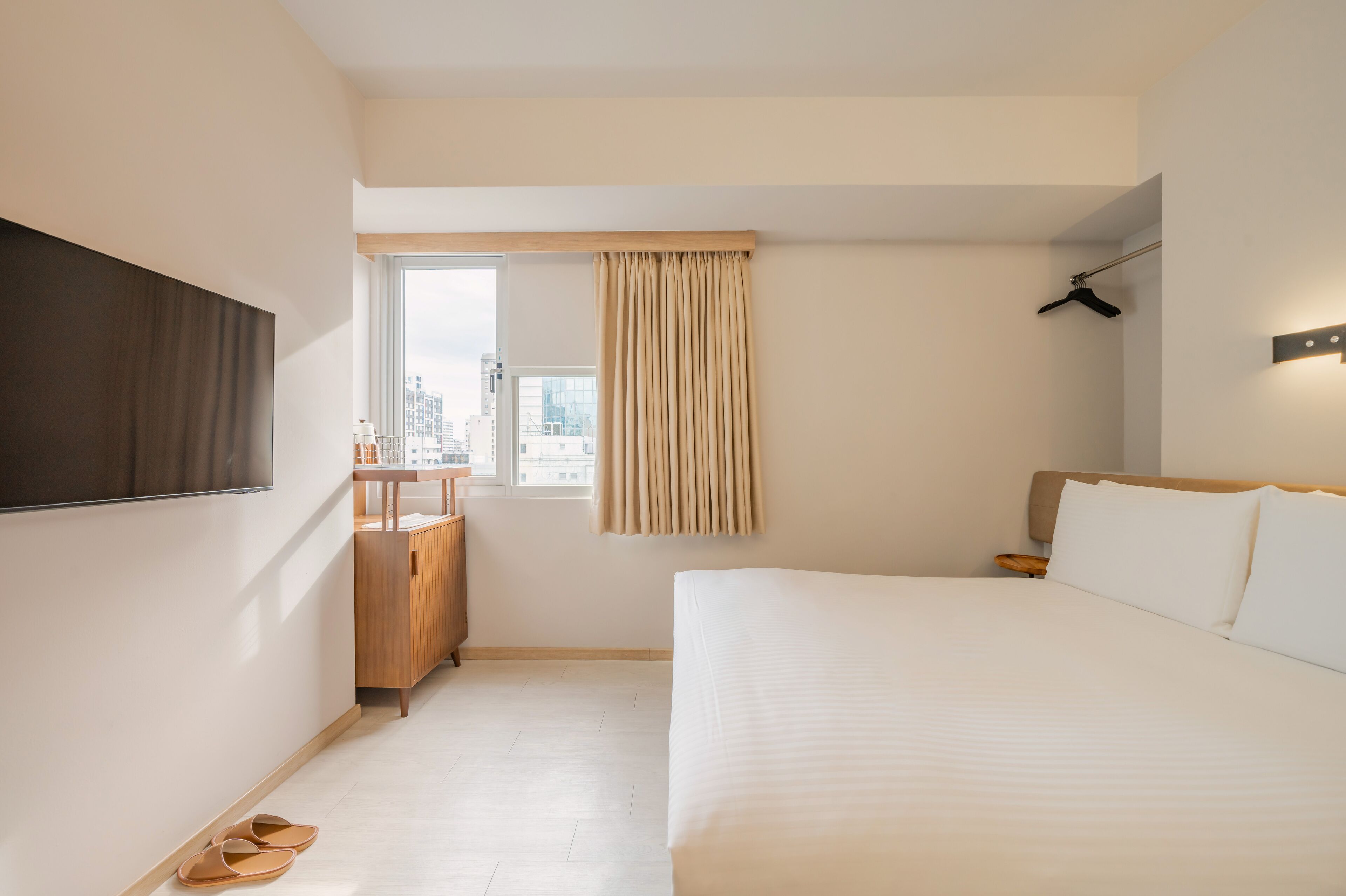 A modern hotel room with a large bed, a TV mounted on the wall, a wooden cabinet, and a window with curtains.