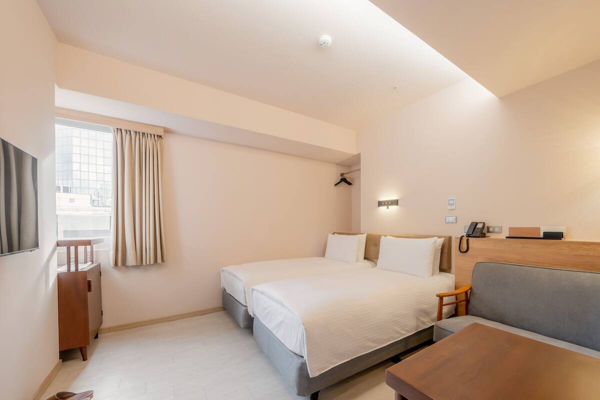 A hotel room with a bed, a sofa, a desk, a TV, a window with curtains, and a ceiling-mounted security camera.