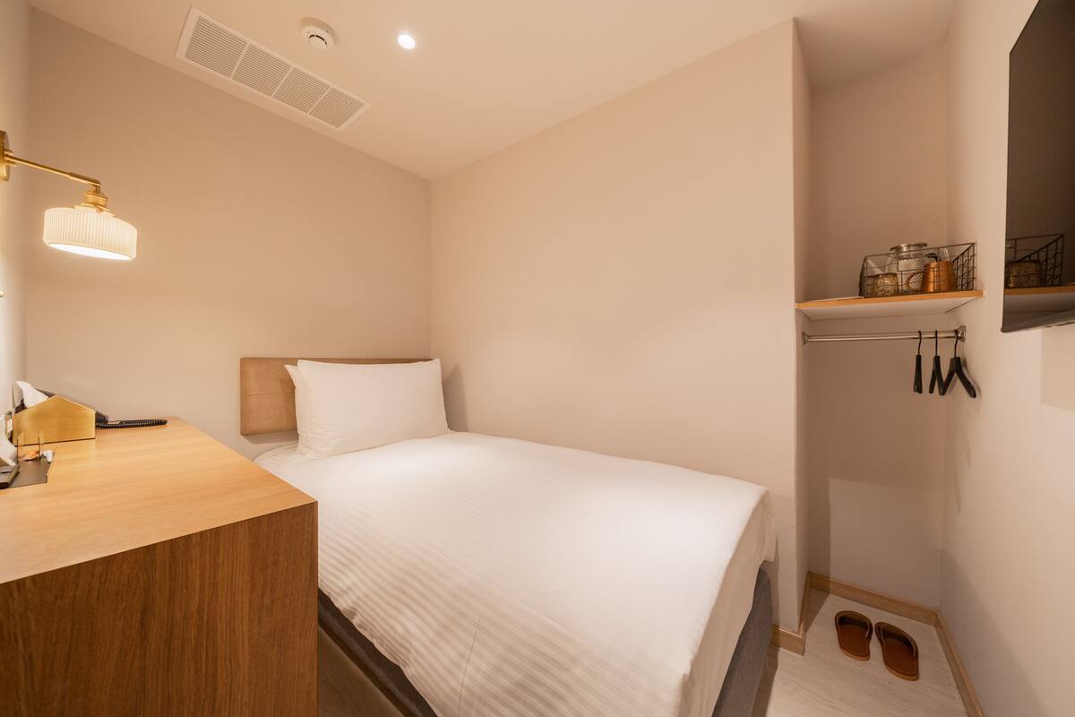 A hotel room with a bed, a wooden desk, a lamp, a shelf with items, and a shoe rack.