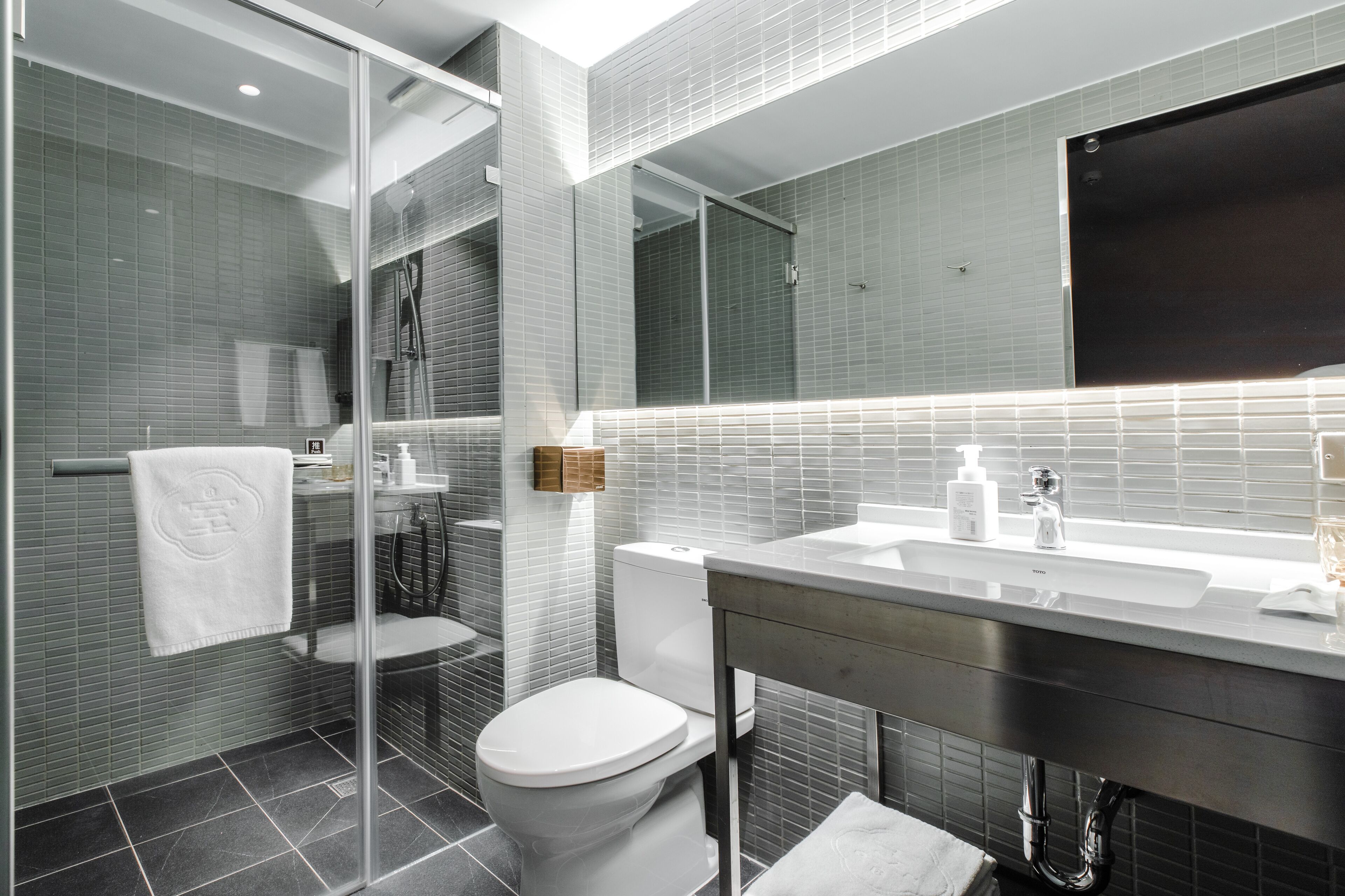A modern bathroom with a glass shower enclosure, a white toilet, a large mirror, and a countertop with a sink and toiletries.