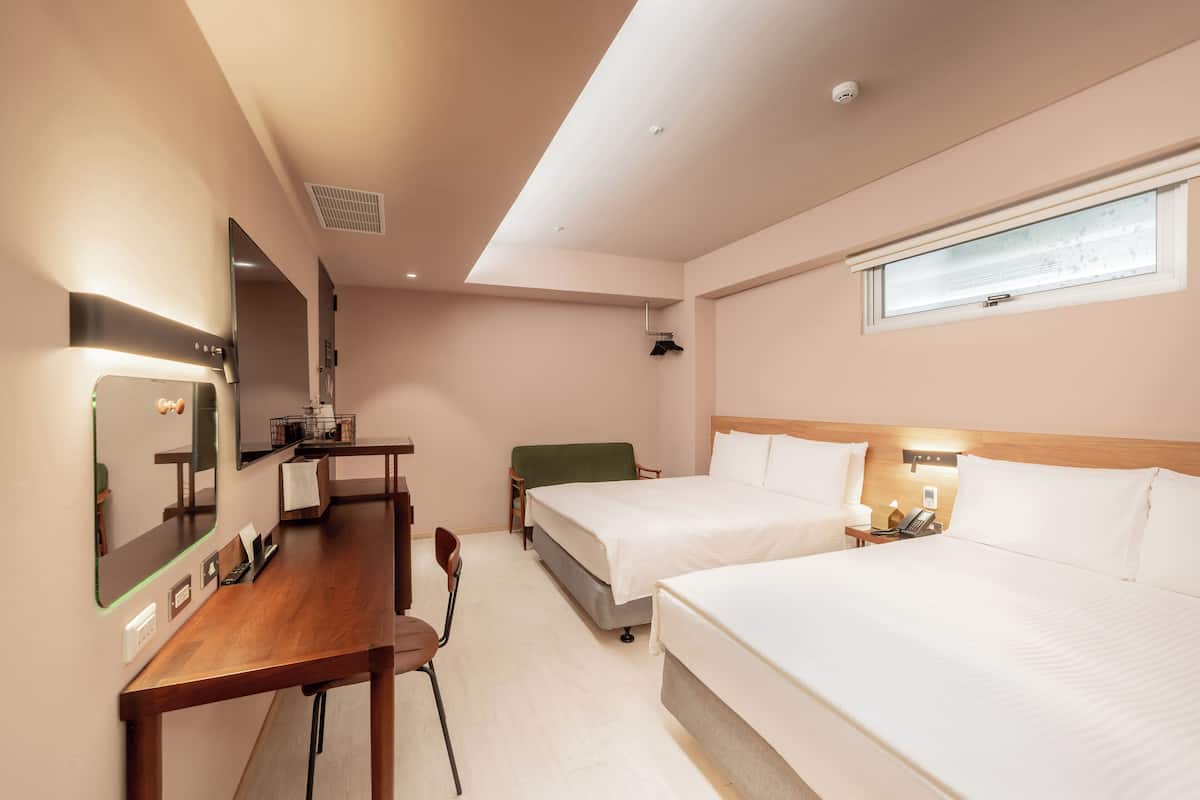 A hotel room with two beds, a desk, a chair, a mirror, a window, and a bedside table with a lamp.
