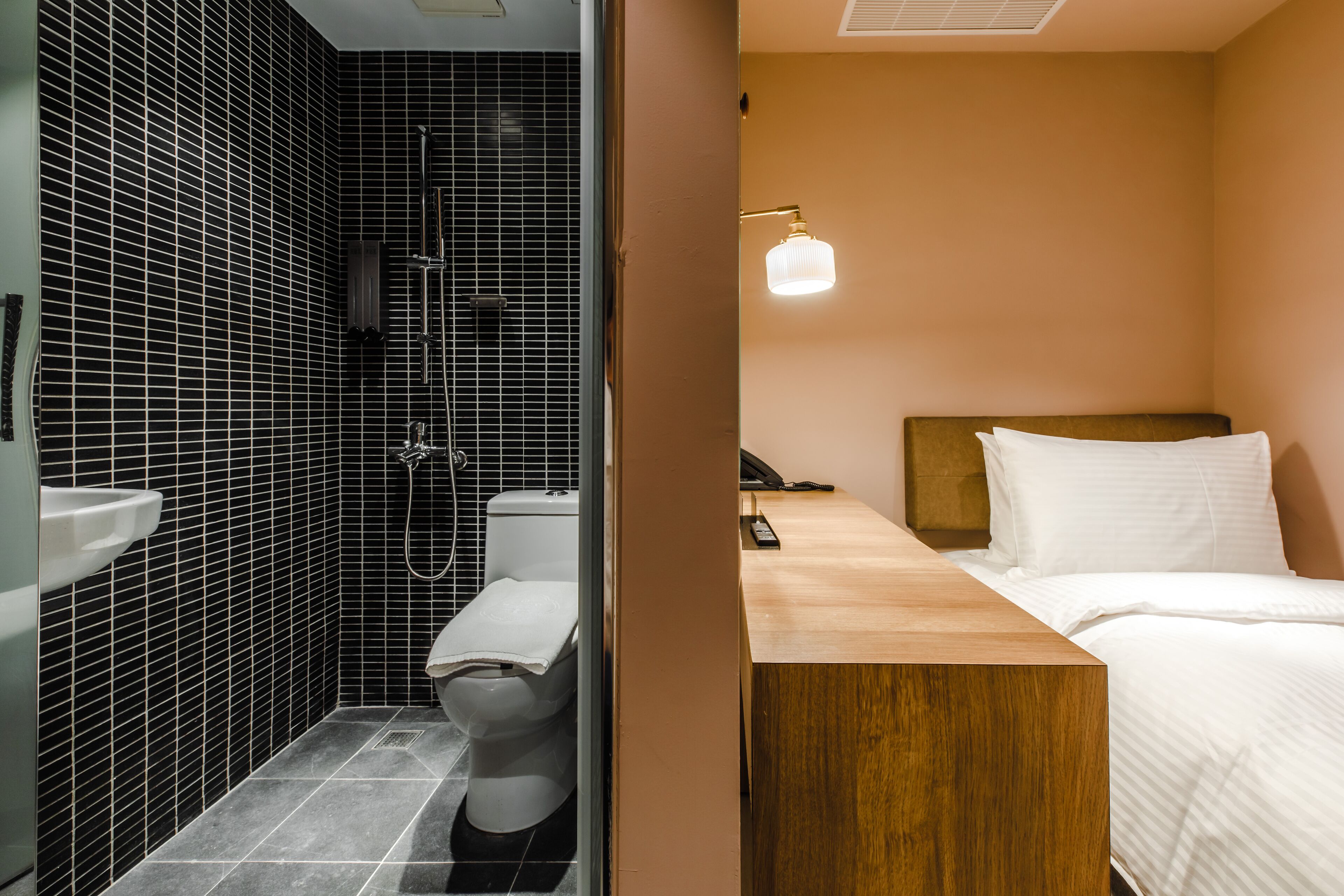 A hotel room with a wooden bedside table, a bed with white bedding, a shower area with black tiled walls, and a white sink.