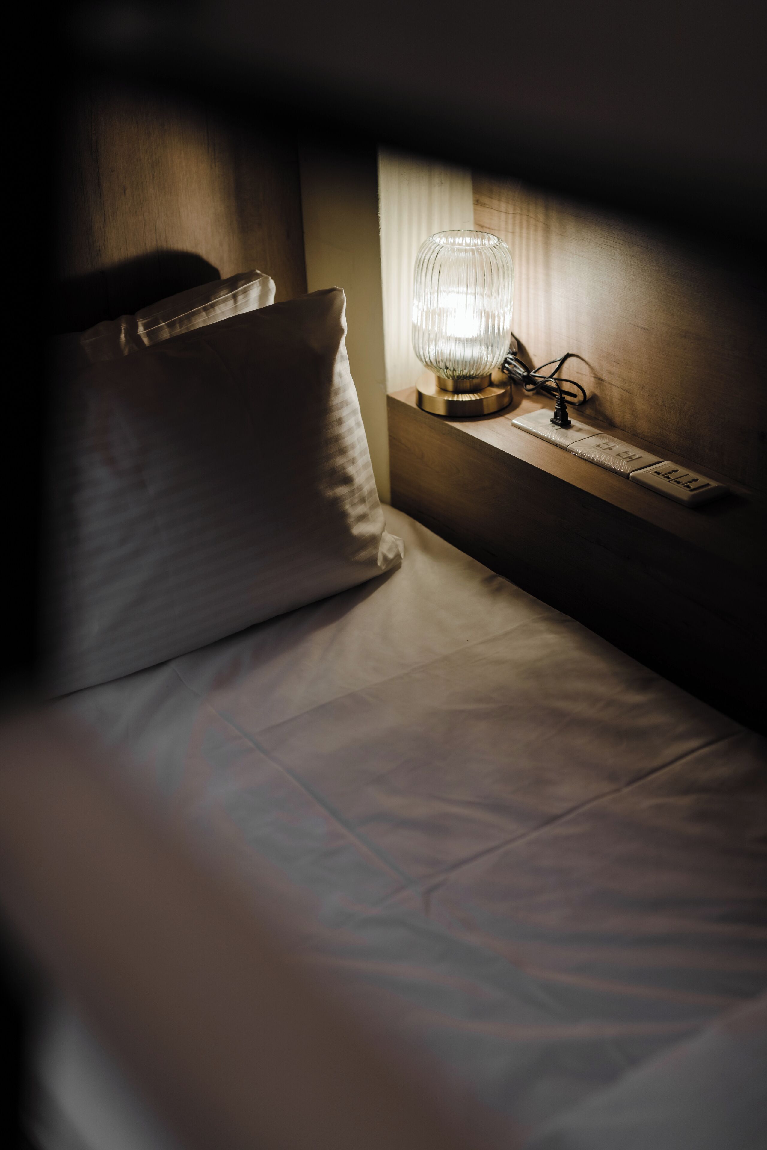 A bed with a pillow, a bedside table with a lamp, and a power outlet.