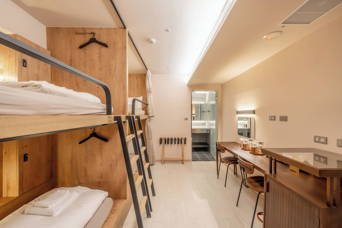 A bunk bed room with a wooden wall, a desk with chairs, and a bathroom visible through a glass door.
