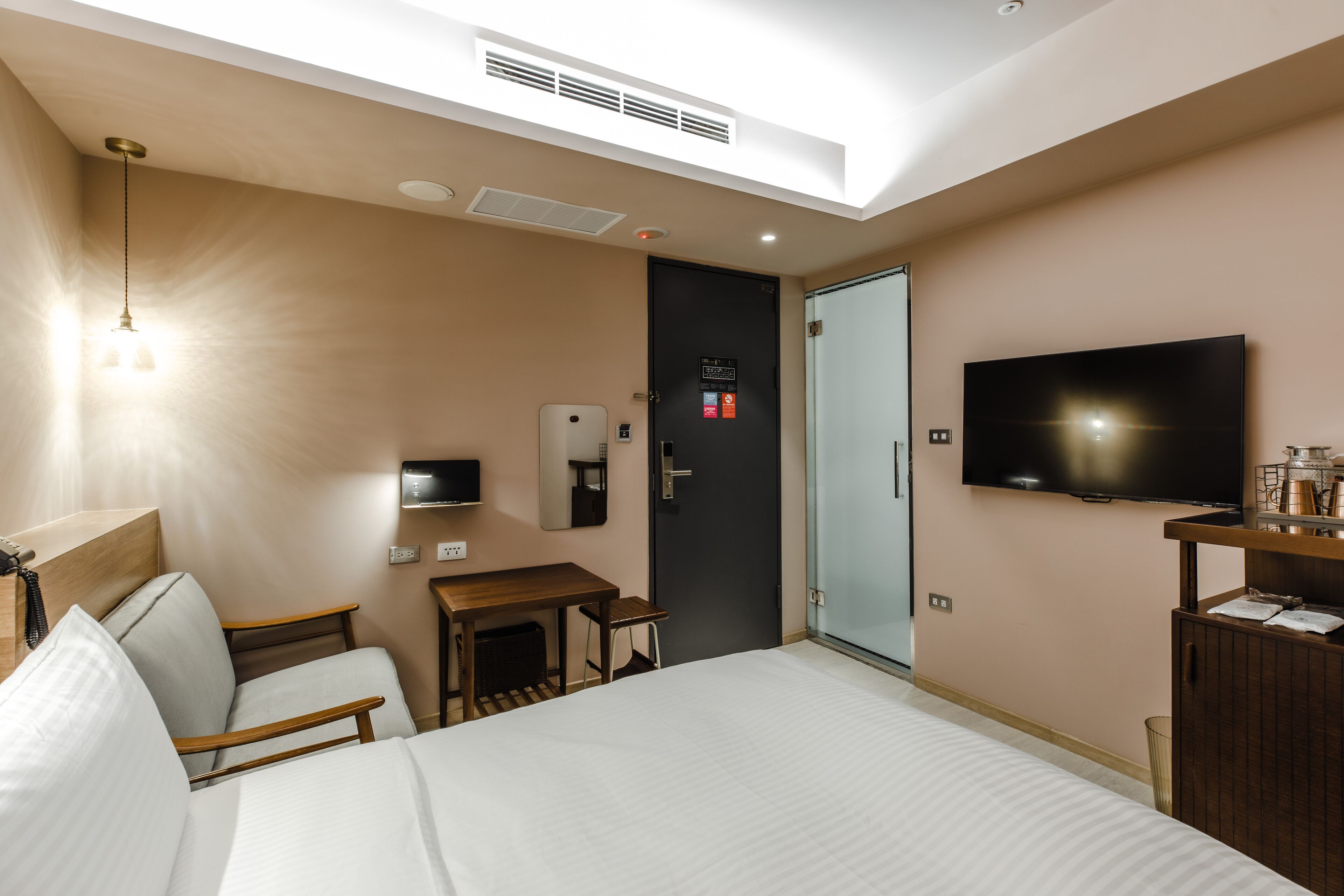A hotel room with a bed, a desk, a chair, a TV, a minibar, and a door with a glass panel.