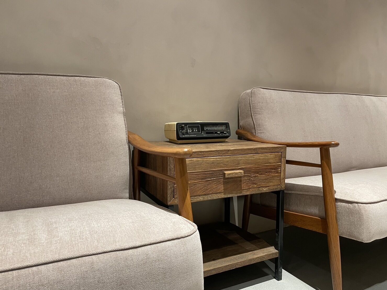 A corner of a room with two beige armchairs, a wooden side table with a radio, and a small wooden drawer.