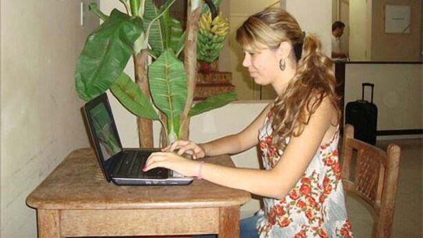 A woman using a laptop at a wooden table with a large plant beside it. - Pousada Mirante das Águas (Aracaju)