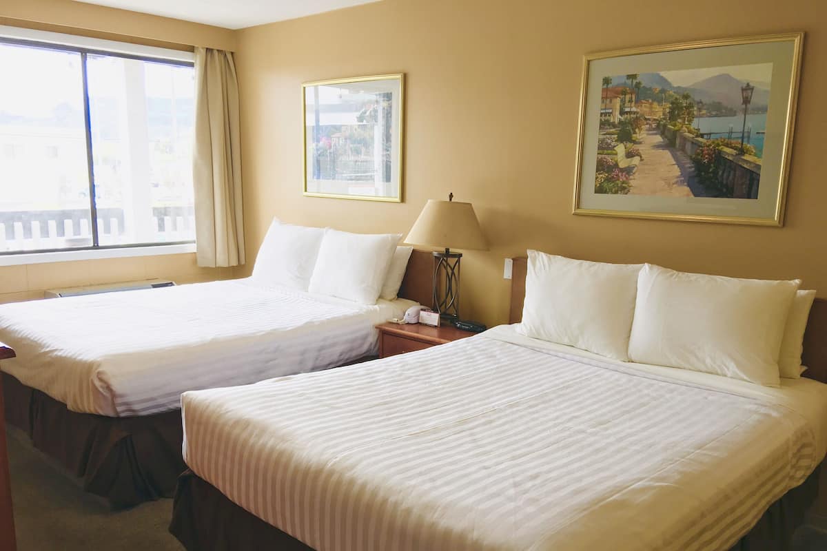 A hotel room with two beds, a window with curtains, a painting on the wall, and a lamp on the nightstand.