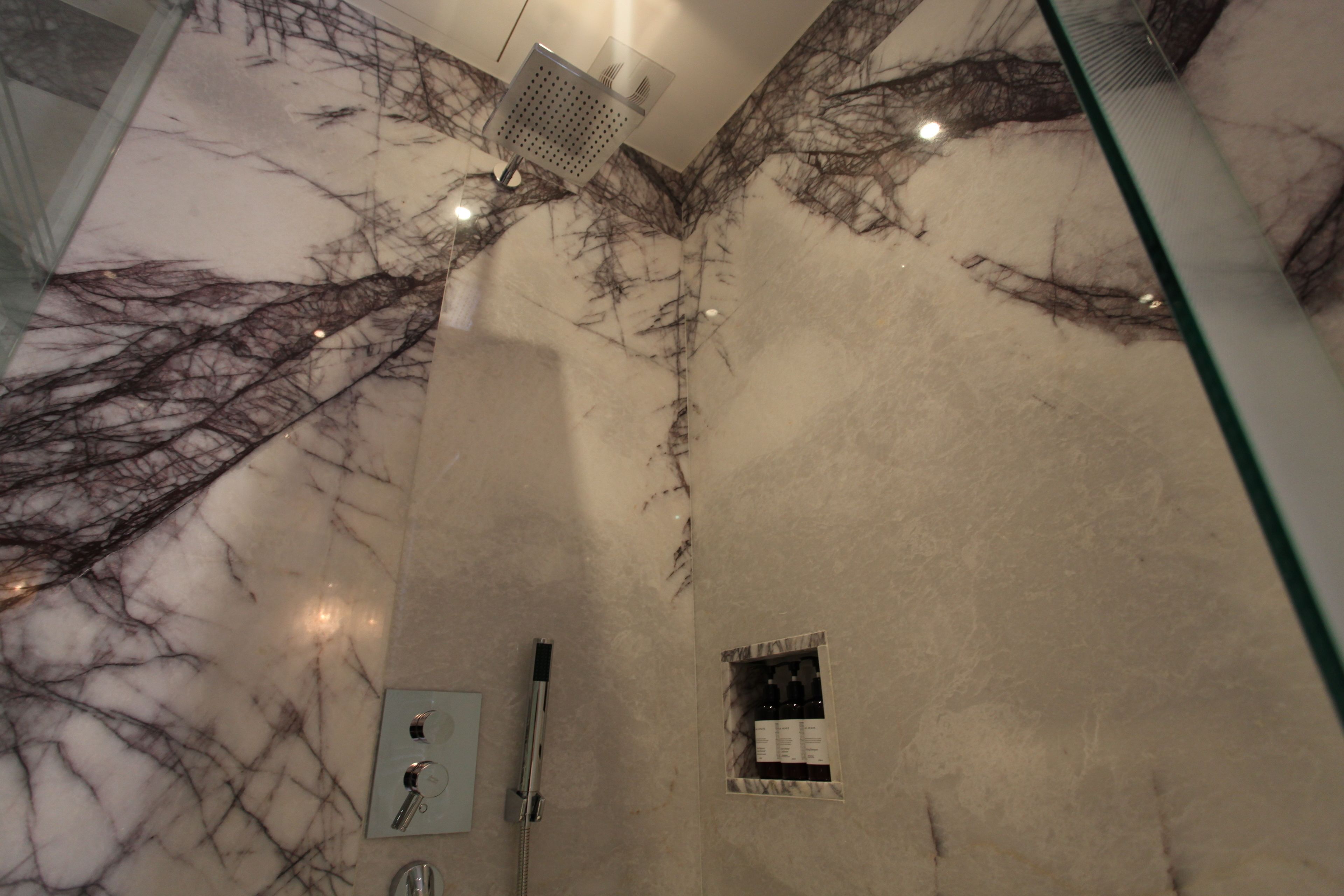 Un baño con pared de mármol, un cabezal de ducha, una ventana pequeña con botellas y un espejo.