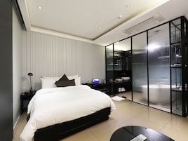 Une chambre moderne avec un grand lit, une table de chevet avec une lampe, une télévision et un dressing fermé par des parois en verre.