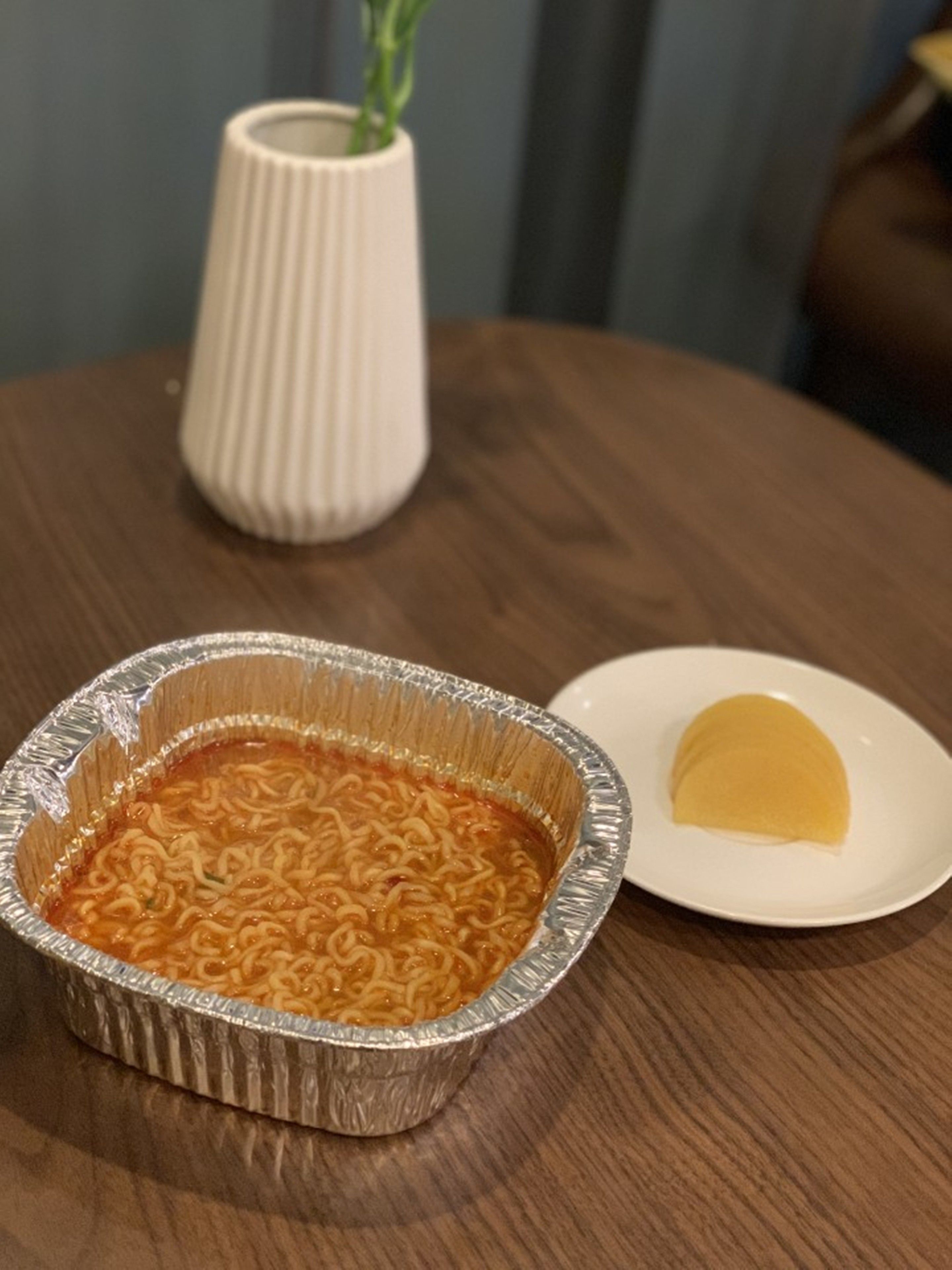 Una mesa con un recipiente de aluminio con fideos ramen y un plato blanco con algo amarillo, posiblemente un postre.