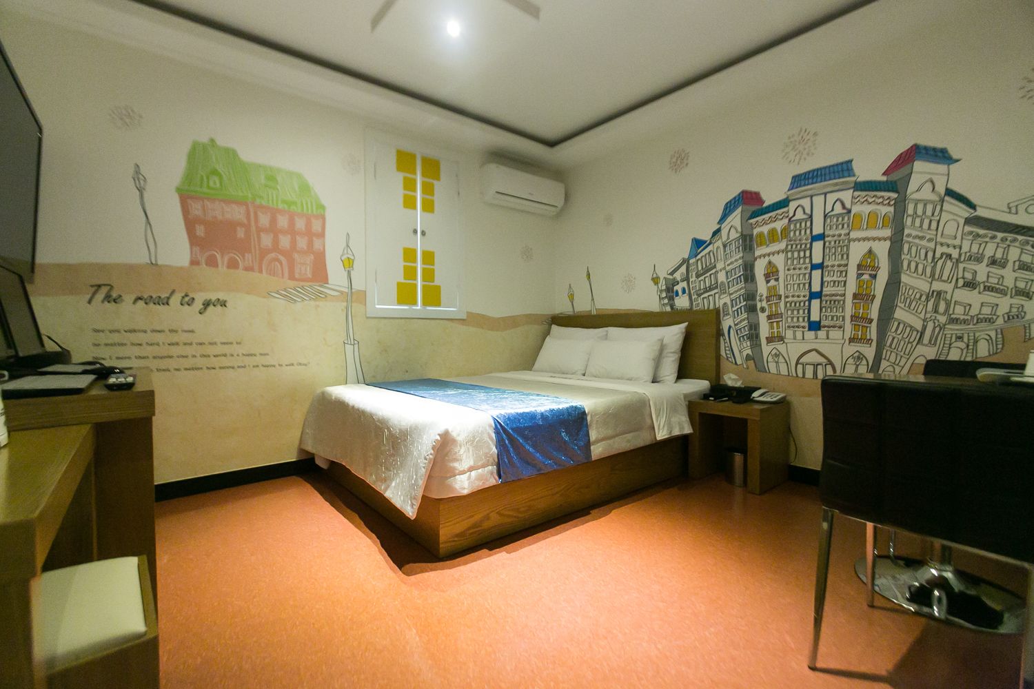 Habitación de hotel con cama, mesita de noche, escritorio y una pared con un mural de un paisaje urbano.