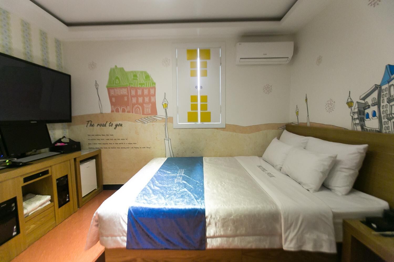 Hotellihuone, jossa on sänky, työpöytä tietokoneella, televisio ja seinä, joka on koristeltu kaupunkimaisemaa esittävällä seinämaalauksella.