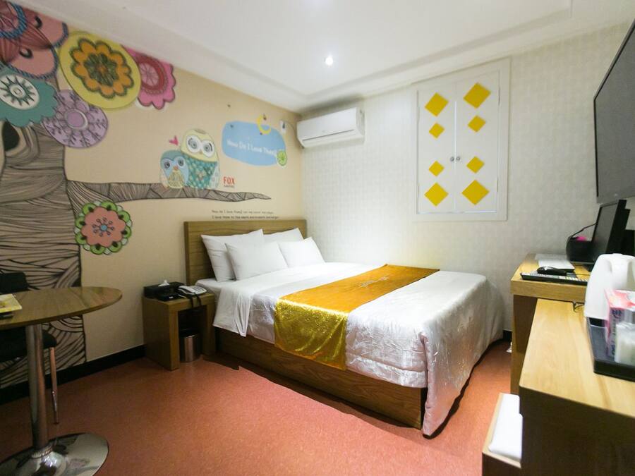A hotel room with a bed, a desk, a chair, a table, and a wall with colorful decorations.