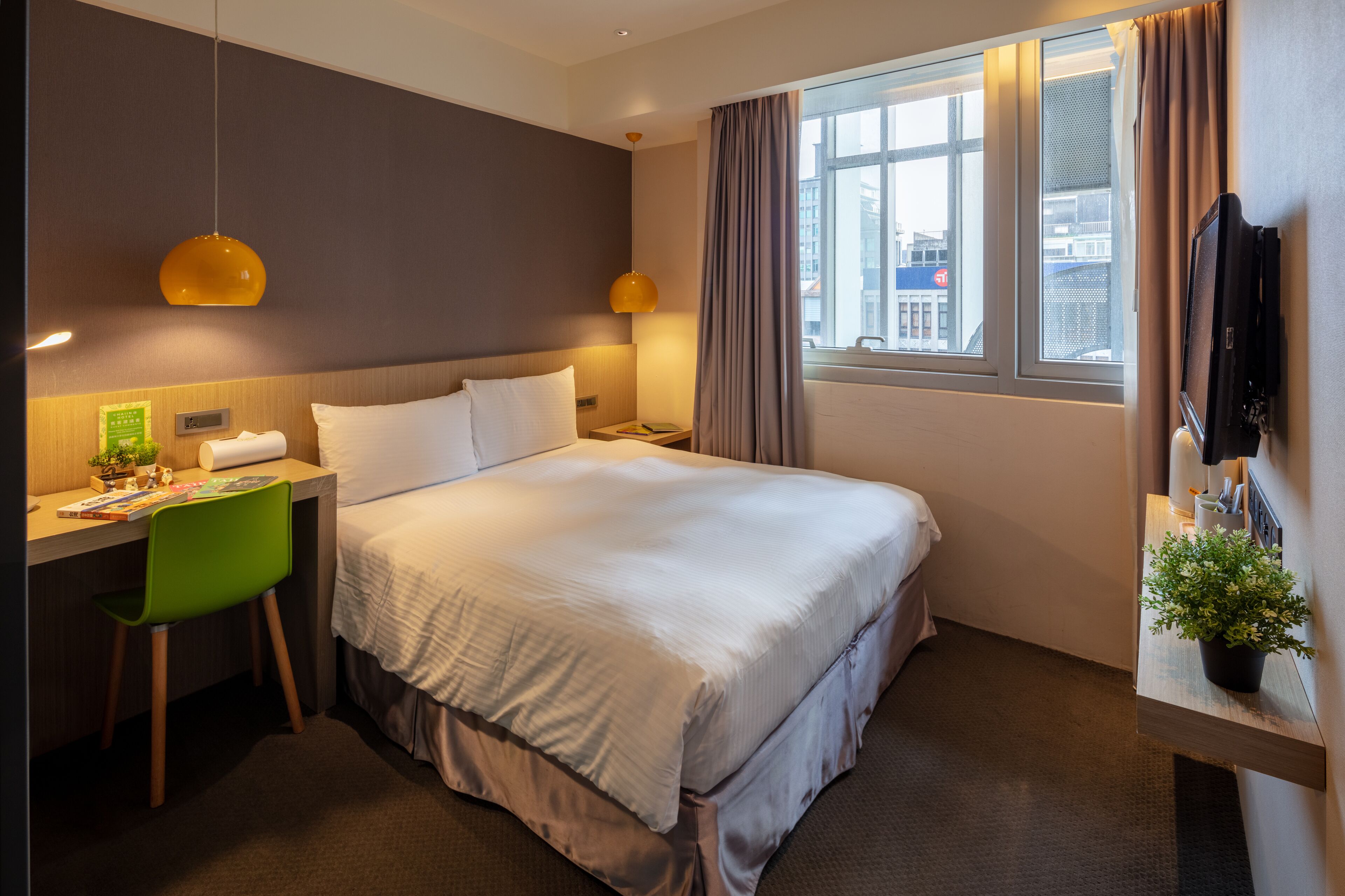 A hotel room with a bed, a desk with a chair, a TV, a window with curtains, and a plant on a side table.