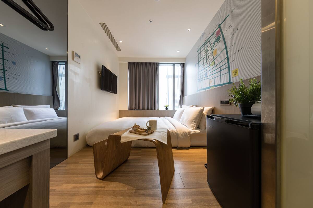 Quarto de hotel moderno com uma cama, uma mesa de madeira, um frigorífico e uma janela grande com cortinas.