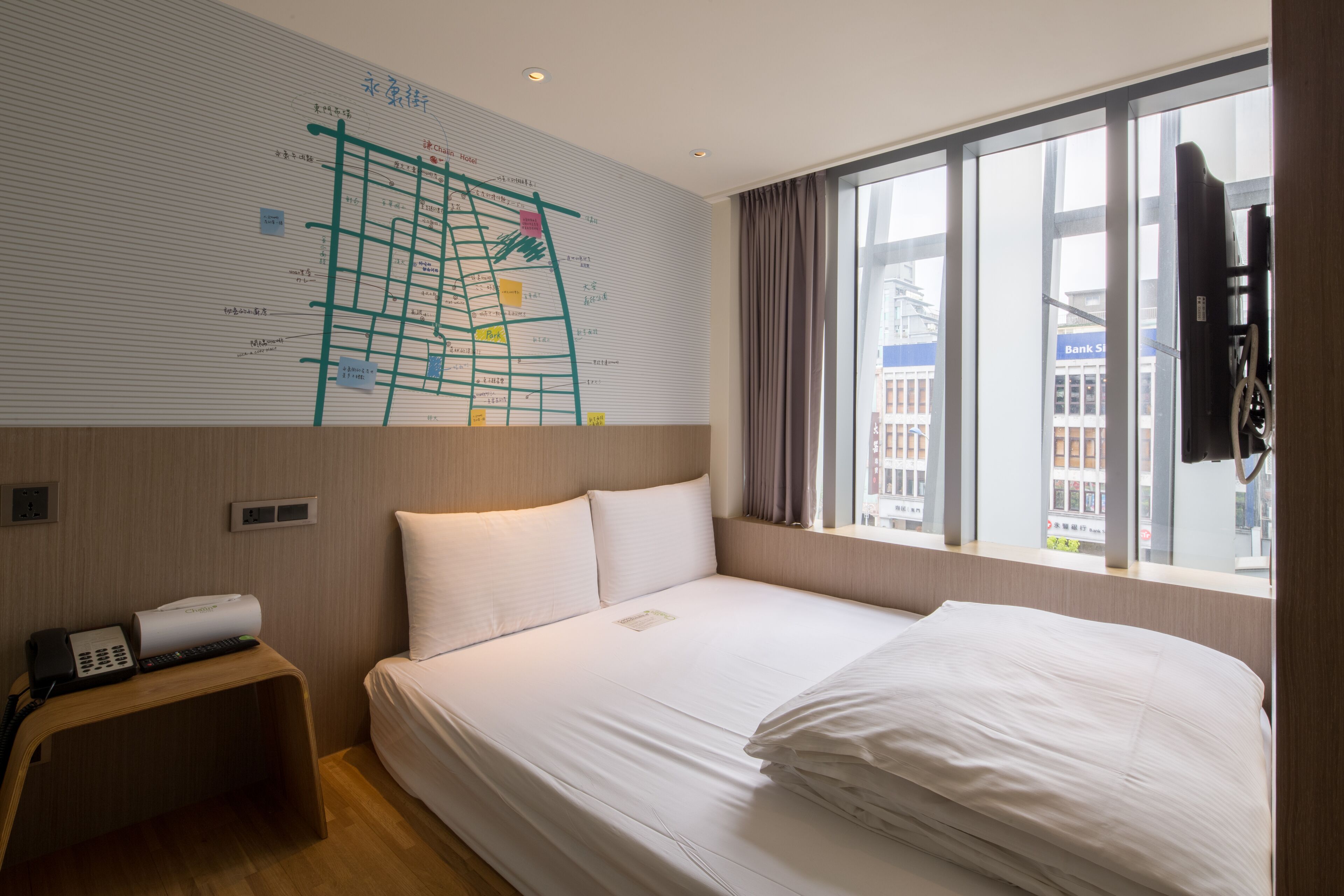 A hotel room with a large bed, a small desk with a phone and remote, a window with curtains, and a wall with a map.