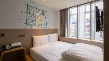 A hotel room with a large bed, a small desk with a phone and remote, a window with curtains, and a wall with a map.