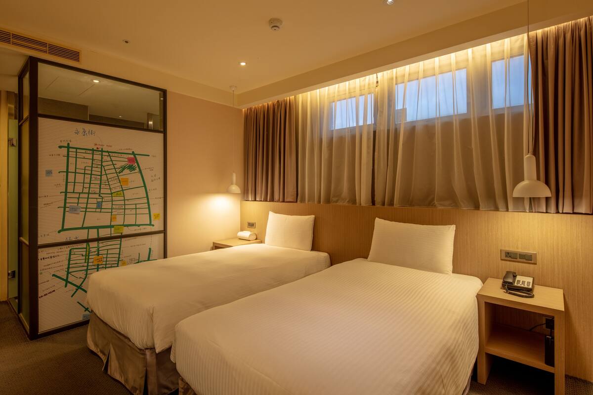 Quarto de hotel com duas camas, uma mesa de cabeceira com telefone, um mapa na parede e uma janela com cortinas.