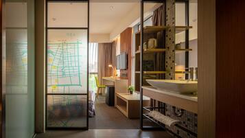 A modern hotel room with a large window, a desk with a chair, a television, a sink, and a mirror.