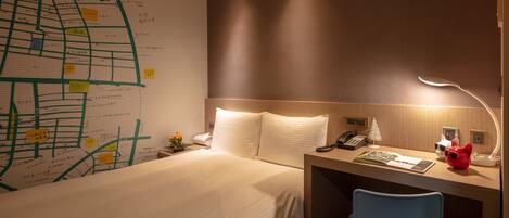 A hotel room with a neatly made bed, a desk with a lamp, a chair, and a wall map.