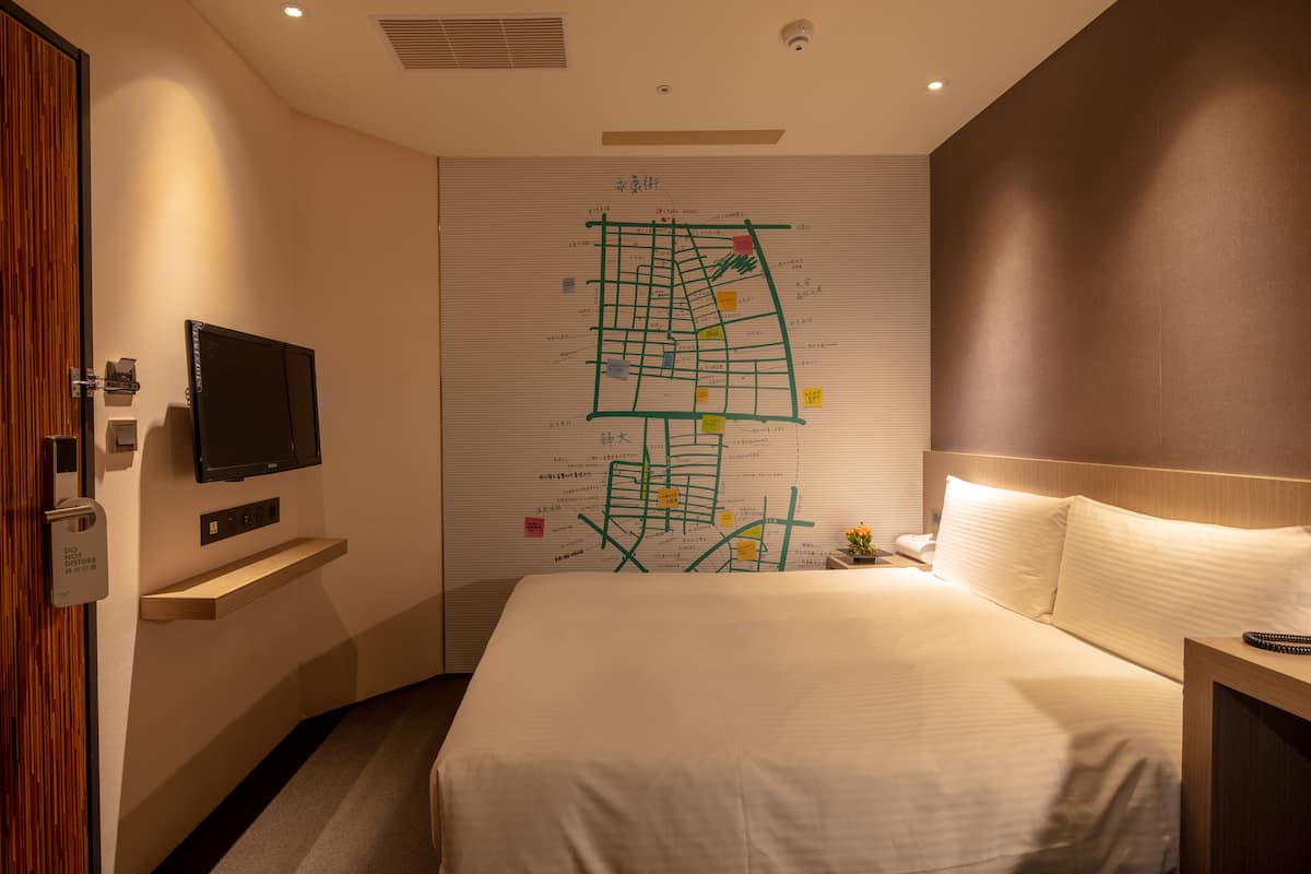 Quarto de hotel com uma cama, uma televisão fixada na parede, um mapa na parede e uma pequena mesa com uma flor.