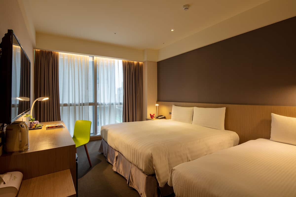 Quarto de hotel com duas camas, uma escrivaninha com abajur, uma cadeira e uma janela grande com cortinas.
