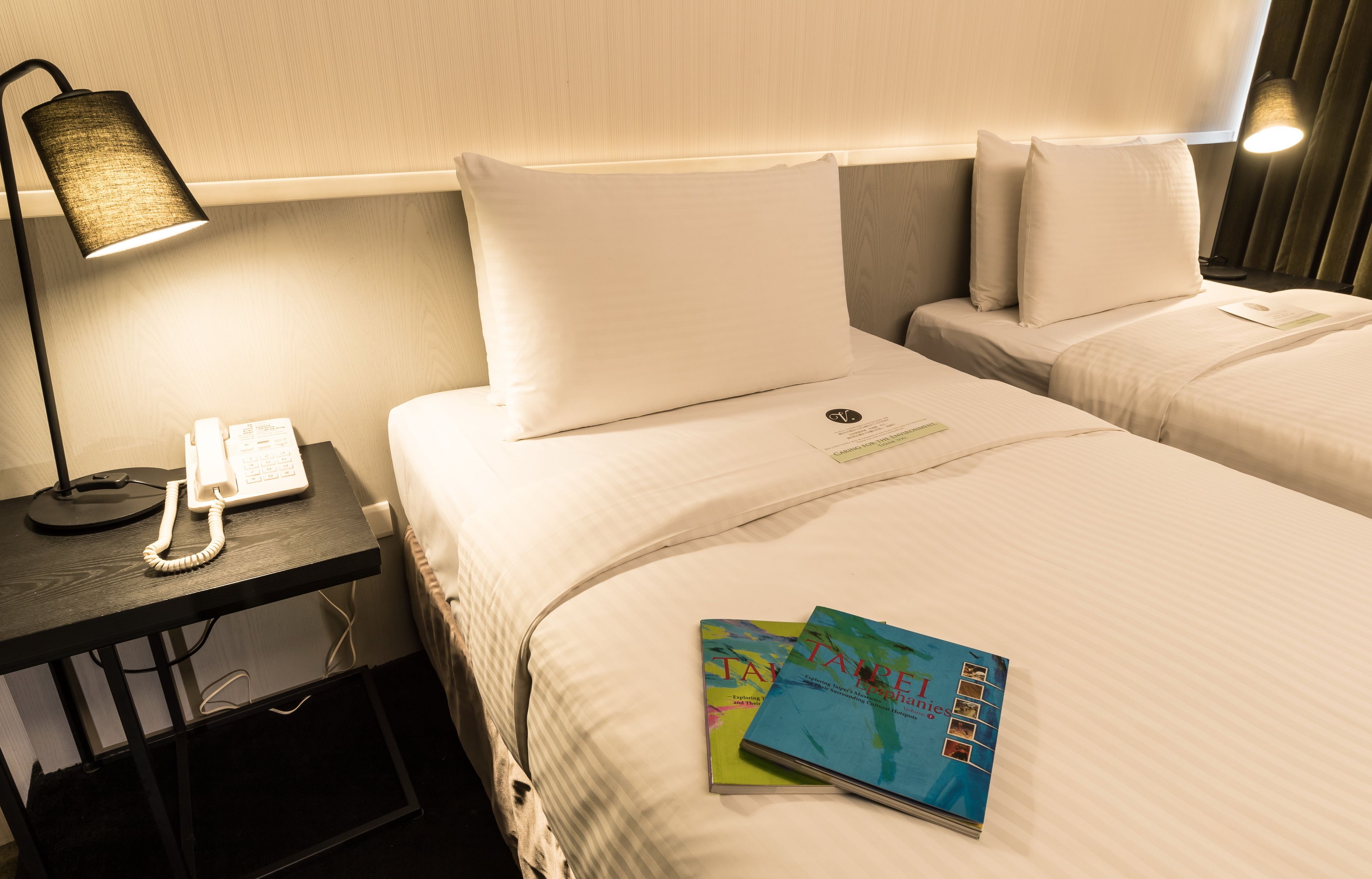 Camera d'albergo con due letti, una scrivania con il telefono e una lampada. Ci sono due libri sul letto.