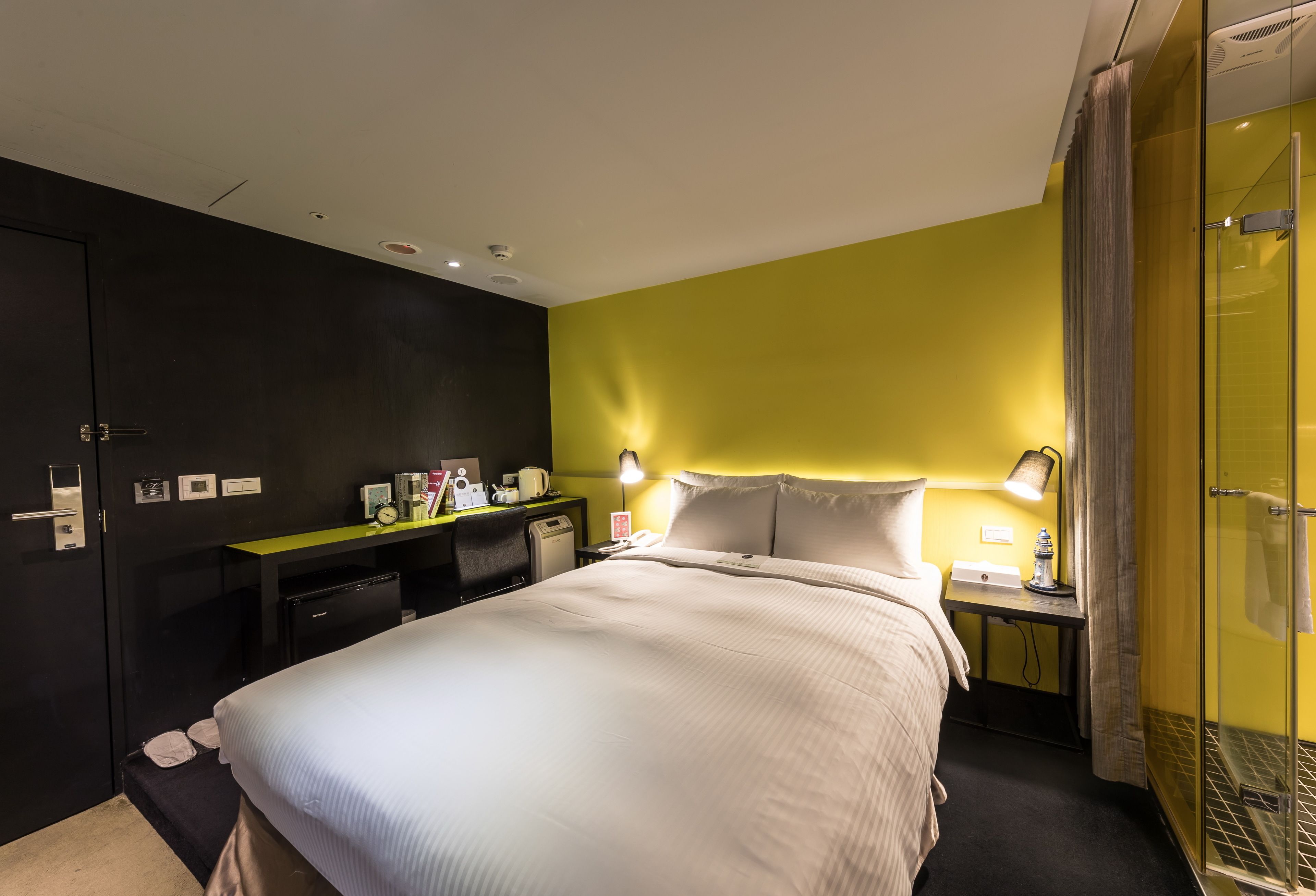 Habitación de hotel con cama, escritorio, silla, mesita de noche, lámpara, espejo, armario y zona de ducha.