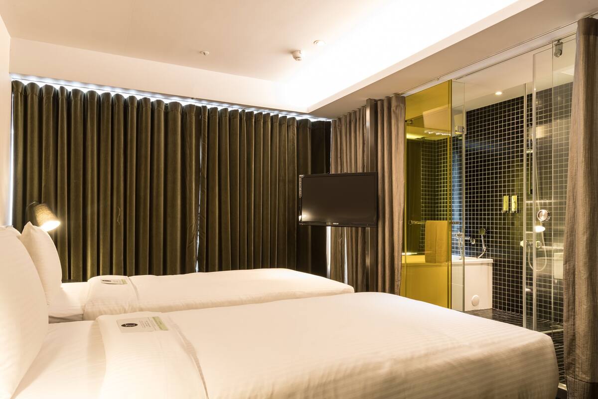 A hotel room with two beds, a flat-screen TV, a glass shower enclosure, and a large window with curtains.