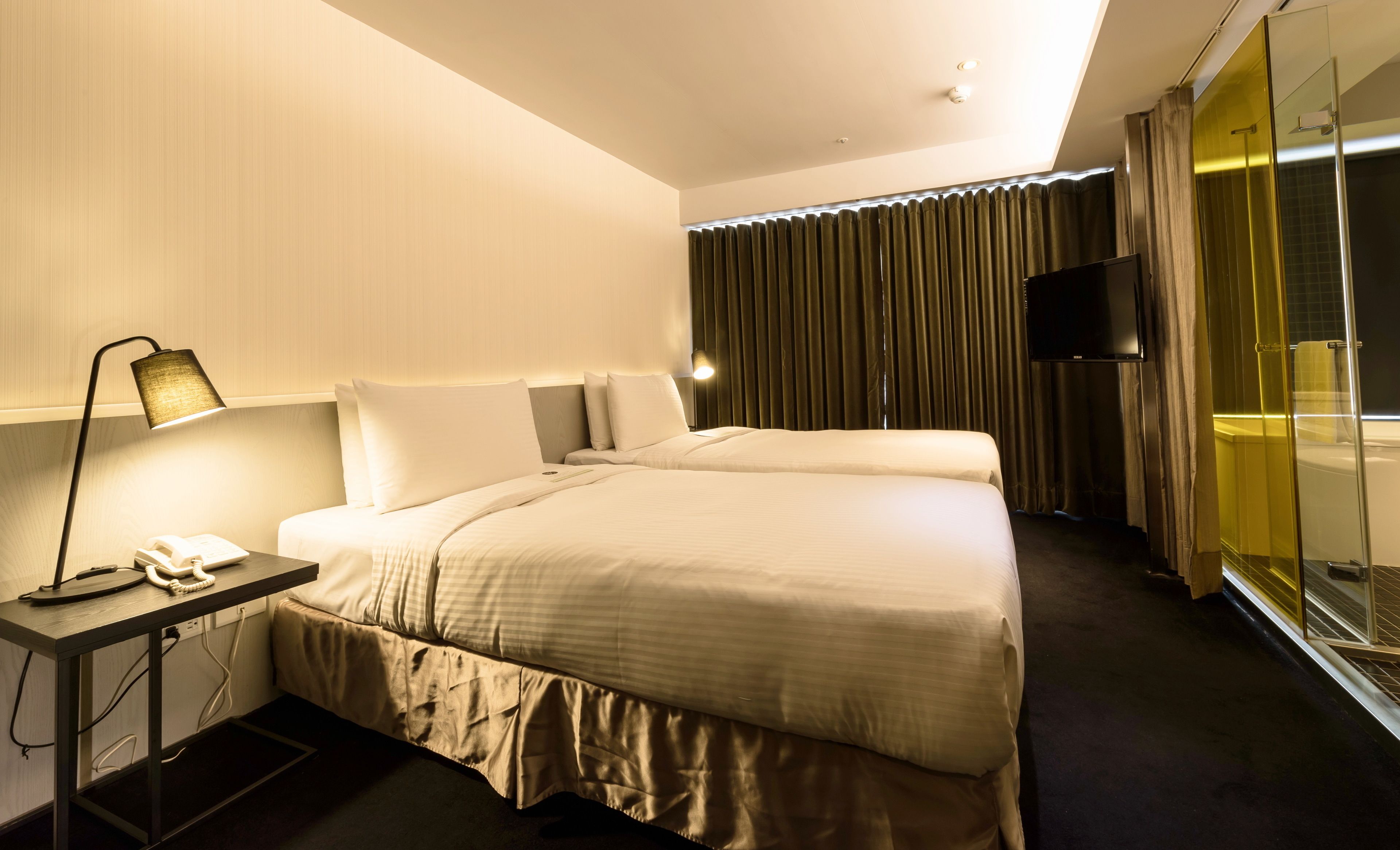 A hotel room with a bed, bedside table with a lamp, a flat-screen TV, and a large window with curtains.