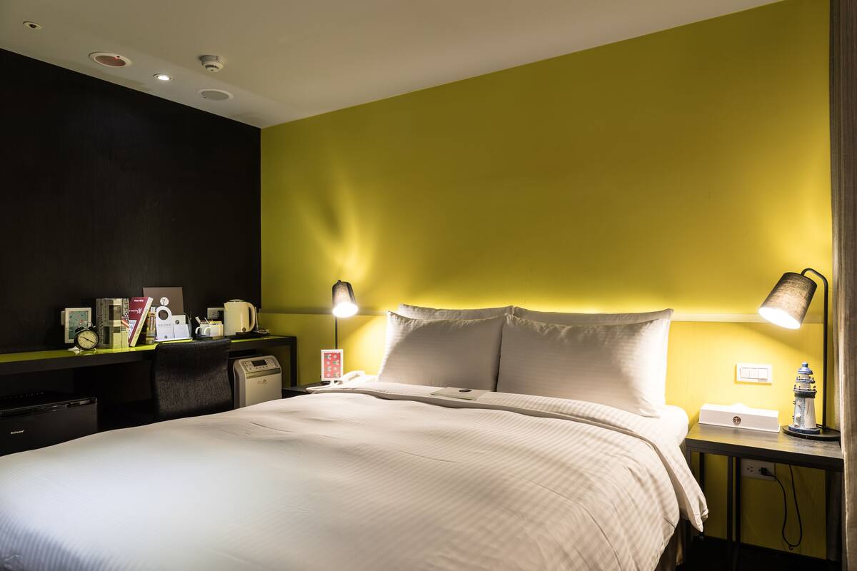 A hotel room with a bed, bedside table, lamp, and a desk with a computer and other items.