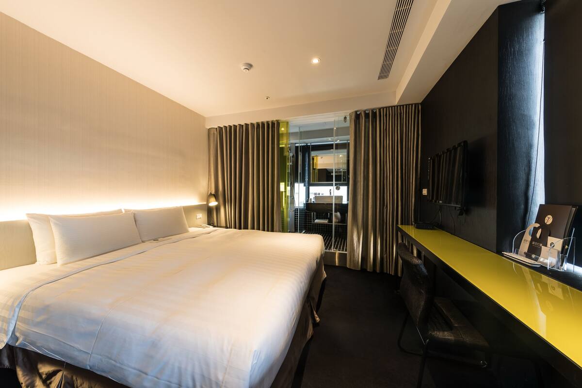 A hotel room with a large bed, a desk with a chair, a mirror, and a view of the bathroom through a glass door.