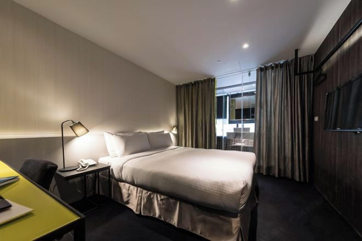 Ein Hotelzimmer mit Bett, Nachttisch, Lampe, Schreibtisch und Blick durch ein mit Vorhängen drapiertes Fenster nach draußen.