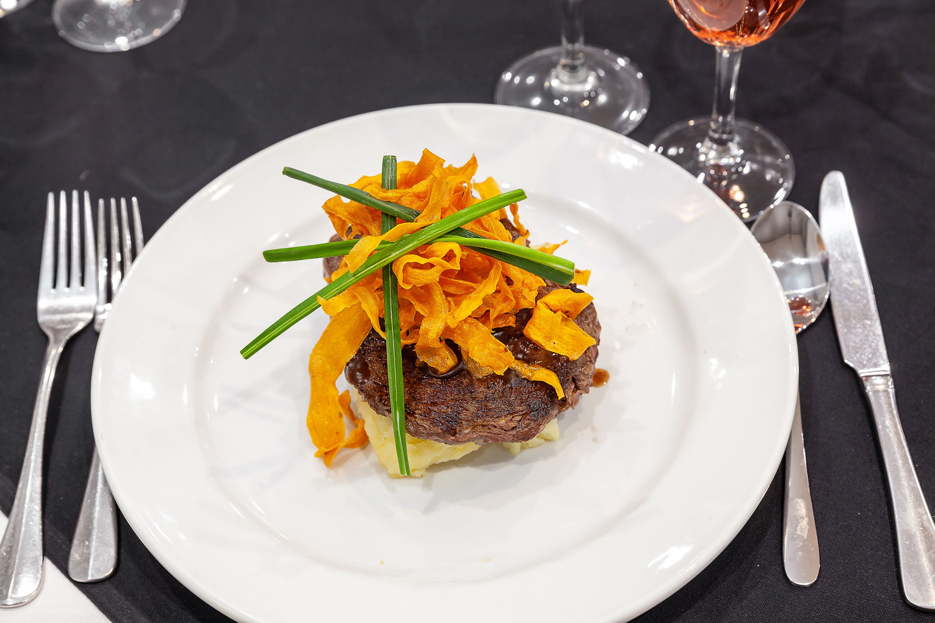 Um prato com bife coberto por um enfeite de legumes laranja e cebolinha verde, acompanhado de talheres sobre uma toalha escura.