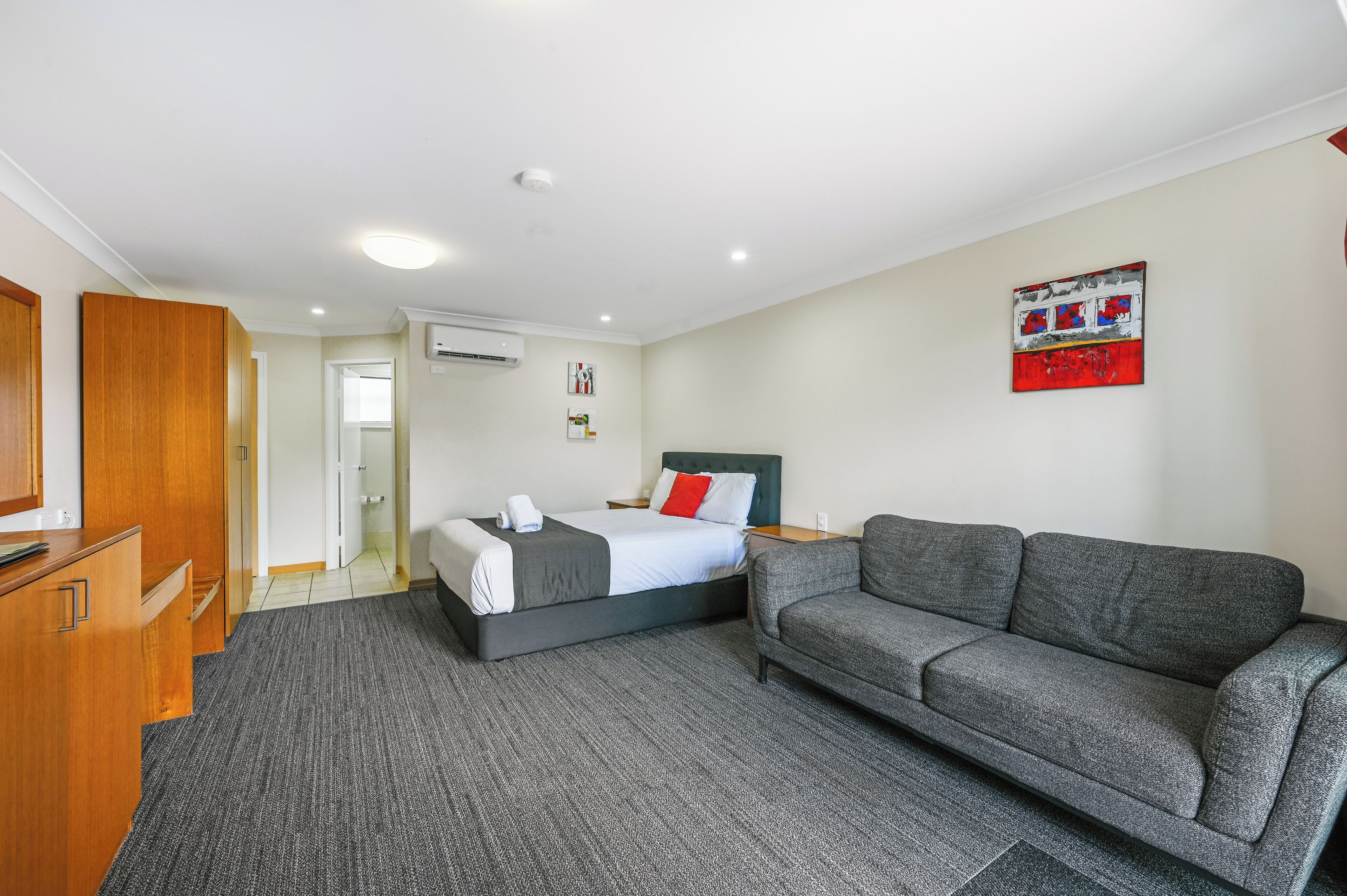 A hotel room with a bed, a grey sofa, a wooden wardrobe, and a painting on the wall.
