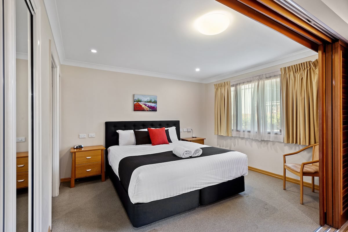 A neatly arranged bedroom with a large bed, a bedside table, a chair, and a window with curtains.