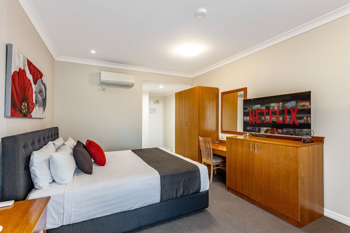 A hotel room with a bed, a desk, a chair, an air conditioning unit, and a television displaying the Netflix logo.