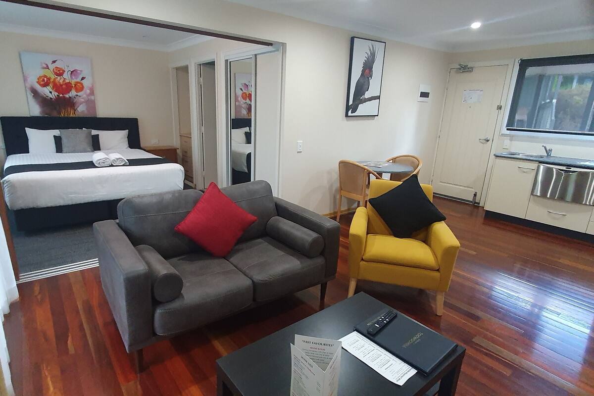 A hotel room with a bed, a grey sofa with a red pillow, a yellow armchair, a small table with a remote control, and a menu on the table.