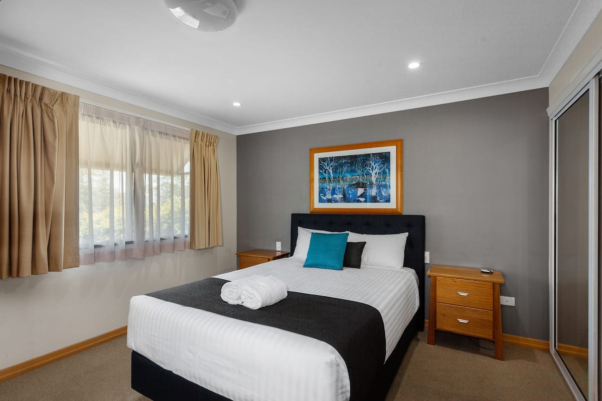 A neatly arranged bedroom with a large bed, a bedside table, a nightstand, and a window with curtains.