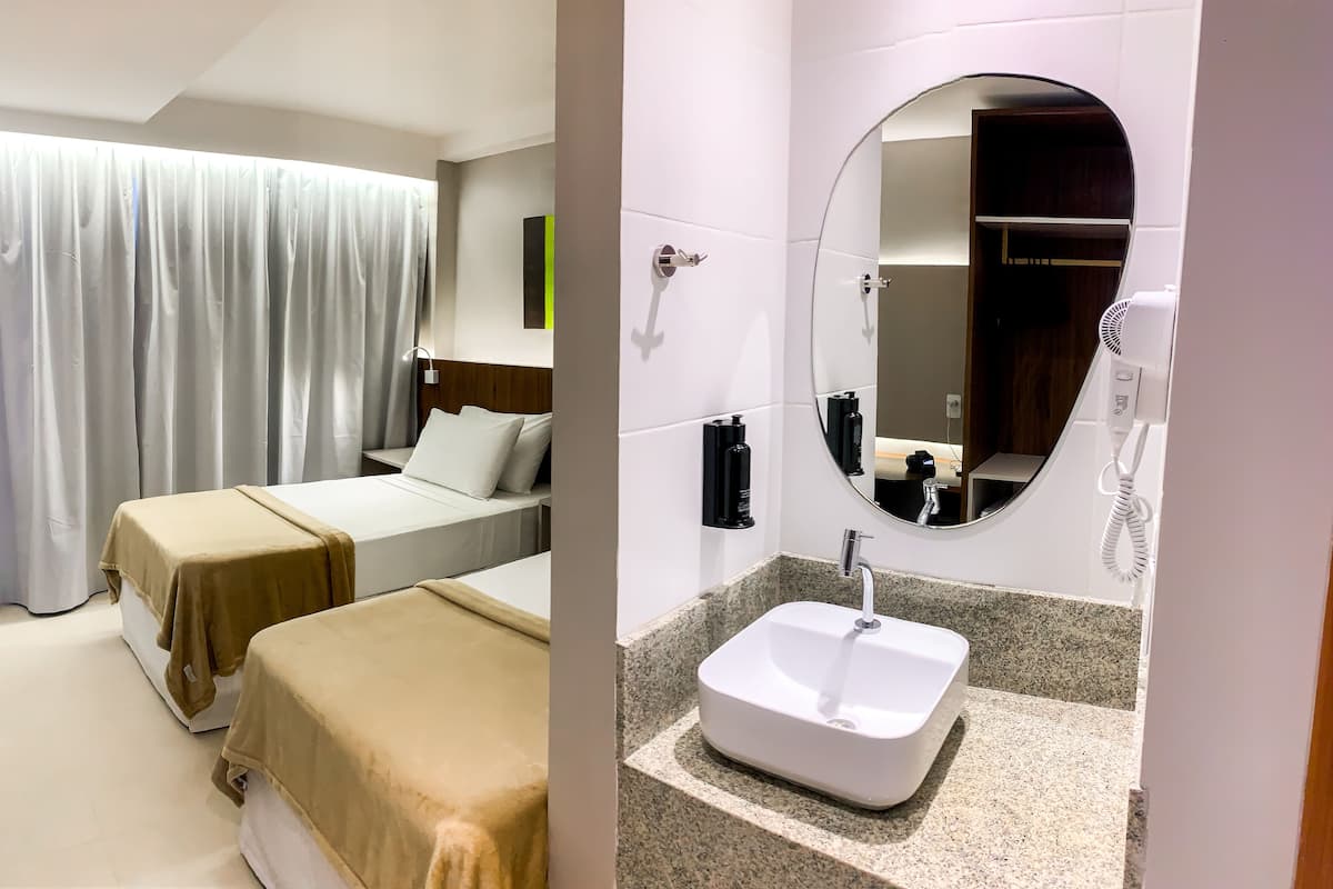 A hotel room with two beds, a large window with curtains, a bedside table with a lamp, and a bathroom with a sink, mirror, and hairdryer.
