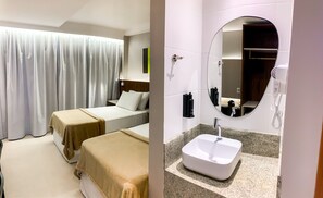 A hotel room with two beds, a large window with curtains, a bedside table with a lamp, and a bathroom with a sink, mirror, and hairdryer. - Smart Hotel (João Pessoa)