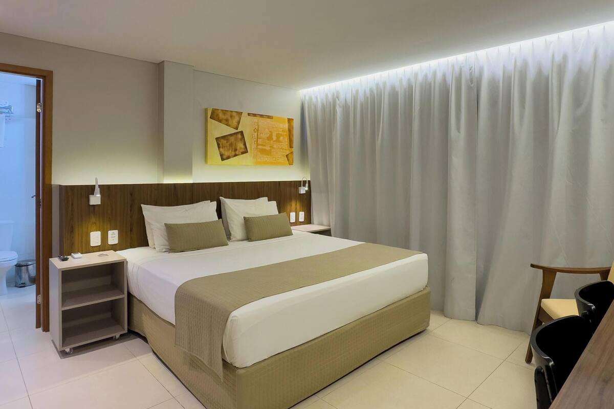 A hotel room with a large bed, bedside tables, a desk with a chair, a television, and a bathroom with a toilet and sink.