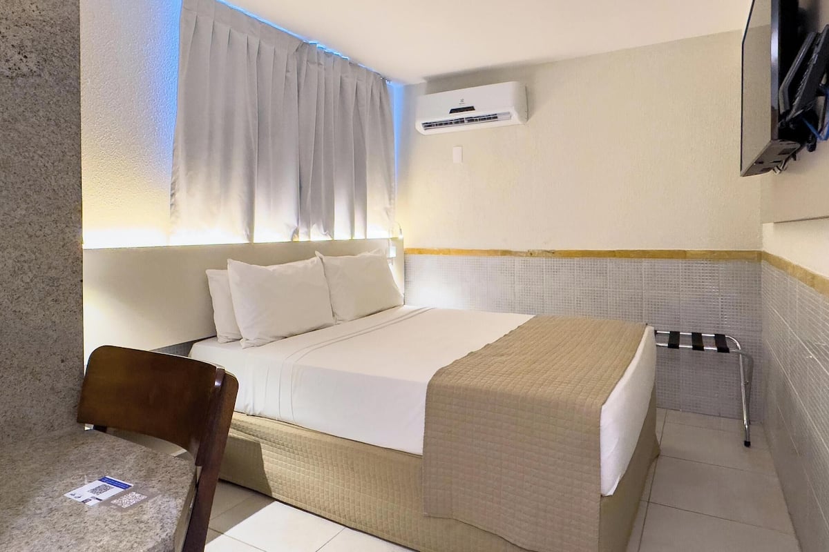 A hotel room with a bed, a chair, a TV mounted on the wall, and an air conditioning unit.