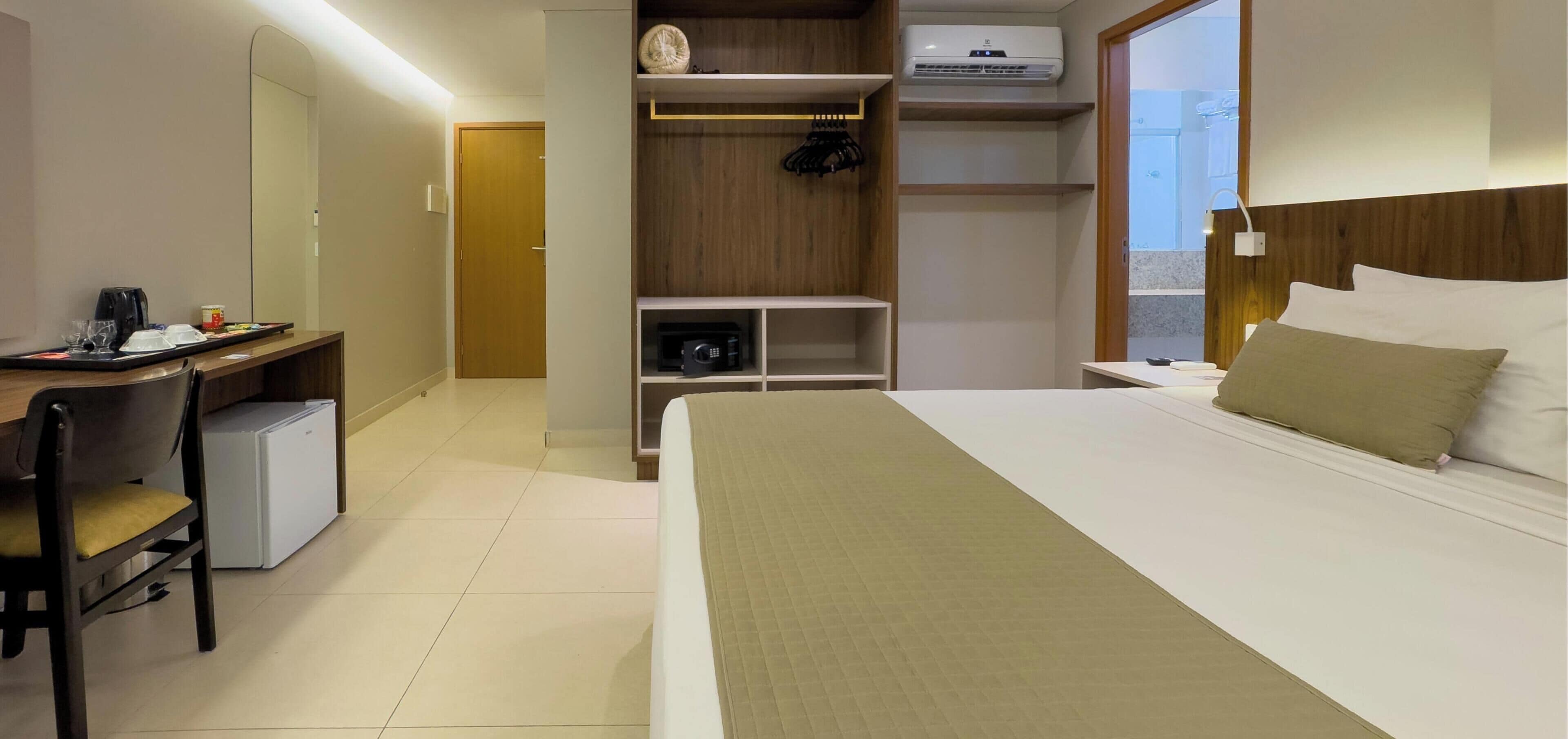 A hotel room with a bed, a desk with a chair, a wardrobe, a microwave, and an air conditioning unit.