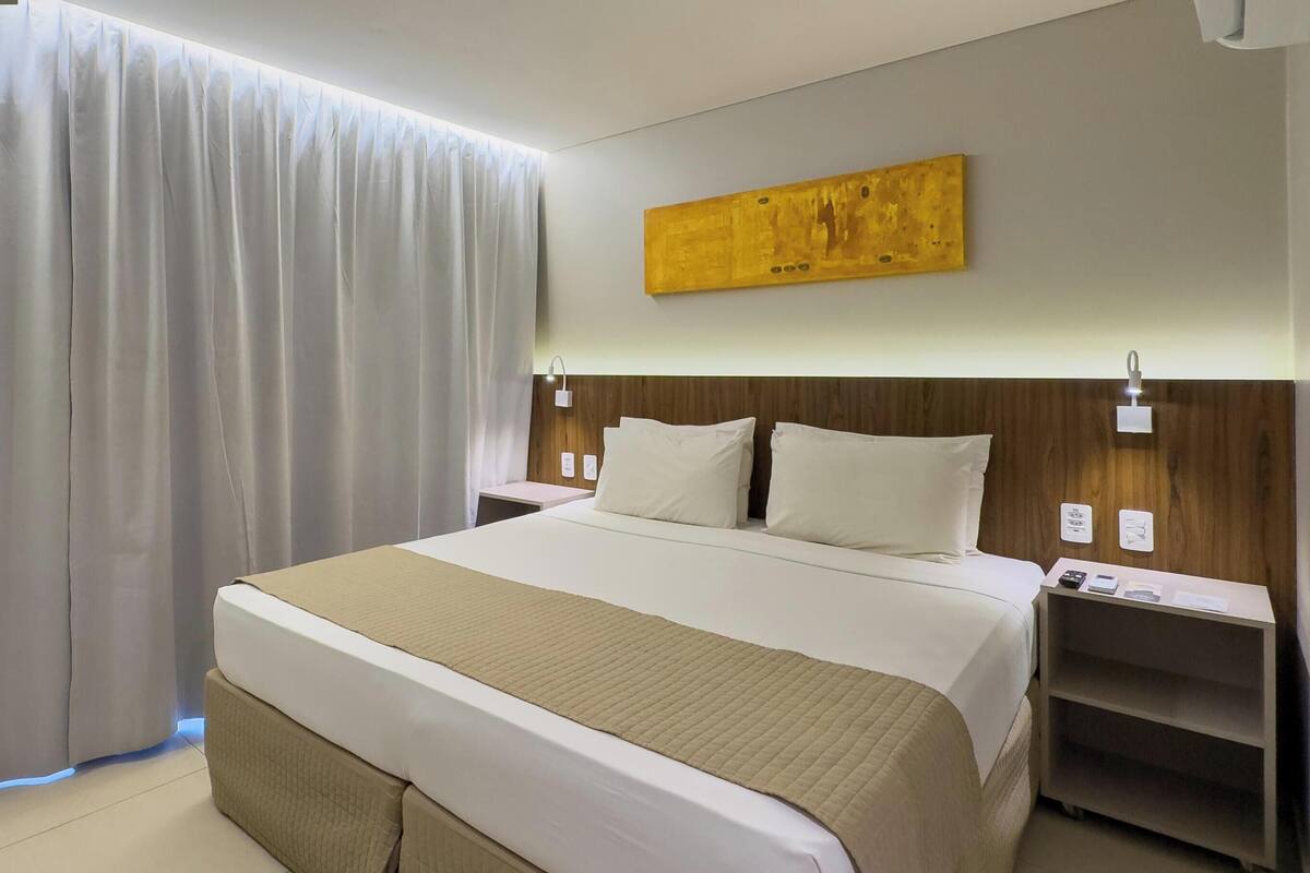 A hotel room with a large bed, a wooden headboard, a bedside table with a lamp, and a wall-mounted artwork.