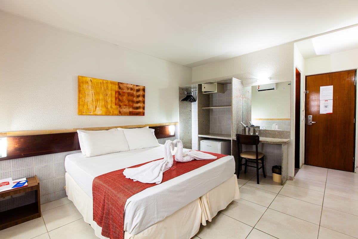 A hotel room with a bed, a desk, a chair, a mirror, a wall-mounted artwork, and a door.
